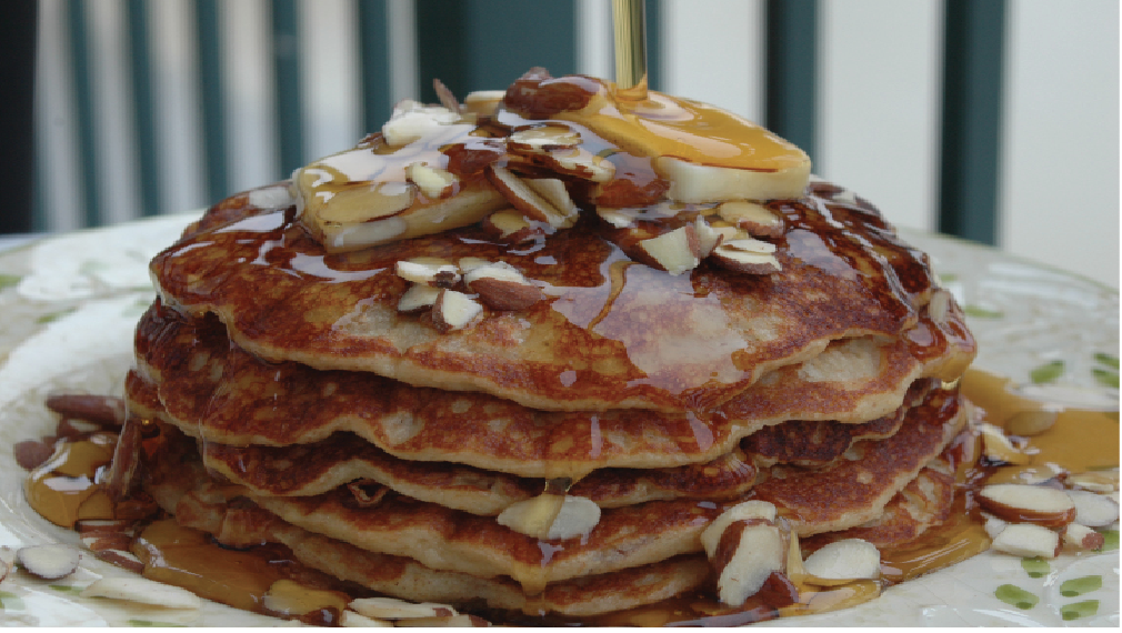 Pancakes sit on a place and syrup drips down ver the top of them covered in sliced almonds